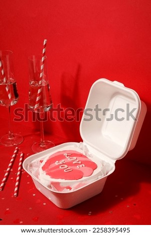 Lunch box with bento cake, glasses of champagne and straws for Valentine's Day on red background