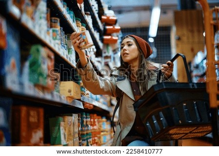 Young woman buying canned food at grocery store. Royalty-Free Stock Photo #2258410777