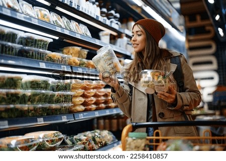 Smiling woman reading label on food package while buying groceries from refrigerated section in supermarket. Royalty-Free Stock Photo #2258410857