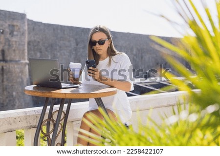 Work in a summer cafe. A young woman with a phone is sitting in front of a laptop. Background of the European old town