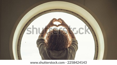 Back view of a woman doing heart sign gesture with hands in a circle window background and light outside. Concept of happiness and healthy freedom lifestyle female people alone. Copy space