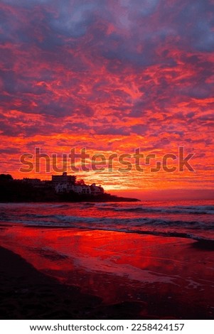 Red sunset at the beach with town in silhouette and dramatic sky with clouds. Amazing sunlight sunrise seascape with waves and houses in background. Concept of travel destination without people