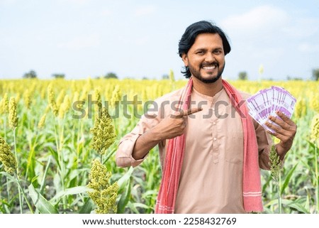 Portrait shot of Happy Indian farmer showing money by pointing finger while looking camera at farmland with copy space - concept of earning, agriculture profit and successful investment Royalty-Free Stock Photo #2258432769
