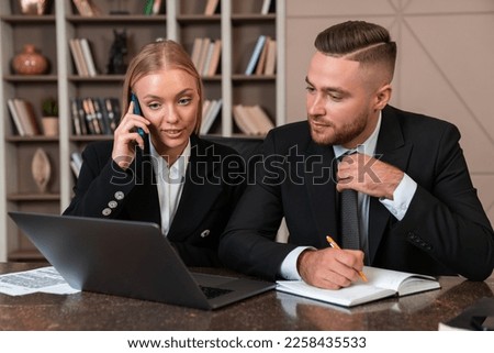 Two business people working together, laptop and notebook on desk. Businesswoman talking on the phone, businessman taking notes. Concept of research and teamwork
