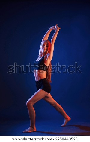 Sportive exercises, standing and stretching the hands. Beautiful muscular woman is indoors in the studio with neon lighting.