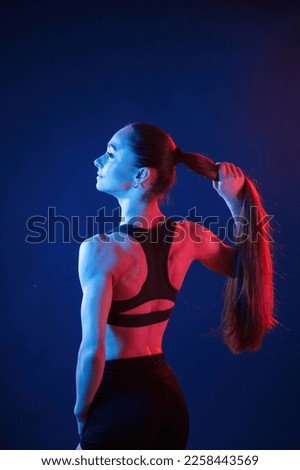 Holding her hair, standing. Beautiful muscular woman is indoors in the studio with neon lighting.