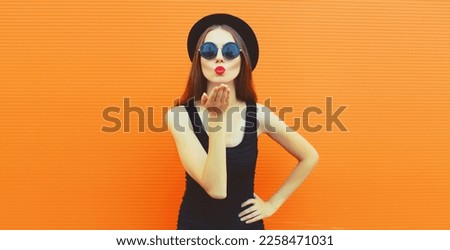 Portrait of stylish young woman model blowing her lips sending sweet air kiss wearing black round hat on orange background