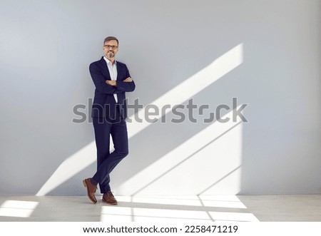 Mature businessman leaning on wall. Full body shot of happy mature business man in stylish dark blue suit and brown leather shoes standing by light gray copy space wall in office. Success concept Royalty-Free Stock Photo #2258471219