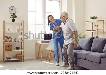 Life of pensioner in modern specialized nursing home. Friendly young female caregiver who cares for elderly man in nursing home helps him get up from sofa. Elderly care concept. Side view.