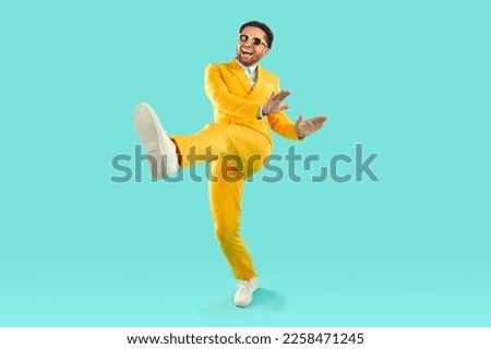 Full length portrait of happy cheerful young man in yellow suit and sunglasses dancing on turquoise background. He is rising one leg and smiling. Banner for advertisement, marketing with copy space.