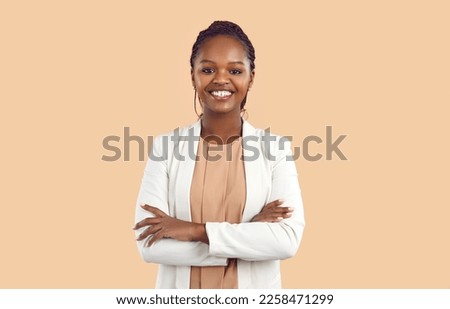 Portrait of young smiling african woman standing with folded arms and looking at camera. Confident stylish business woman in blouse and jacket standing on beige background with copy space. Banner.