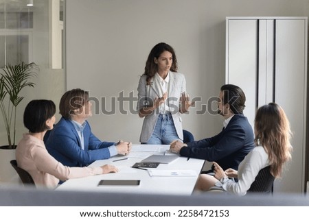 Serious pretty millennial project leader woman talking to employees on meeting, standing at table, explaining tasks, presenting strategy, plan. Female boss mentoring team Royalty-Free Stock Photo #2258472153