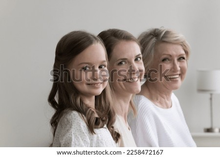 Cheerful pretty daughter kid, young mother and happy mature grandma standing in row, posing at home, looking at camera, smiling. Three female generations family portrait