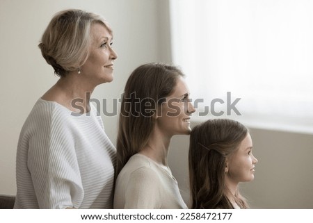 Happy pretty kid girl, young mother and 60s older grandma posing for picture, standing In row, looking at window away. Family side portrait with three female generations