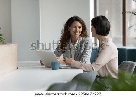 Two positive confident diverse business colleagues women talking at workplace, discussing project documents, at laptop, speaking, smiling, laughing. Mature manager consulting expert Royalty-Free Stock Photo #2258472227