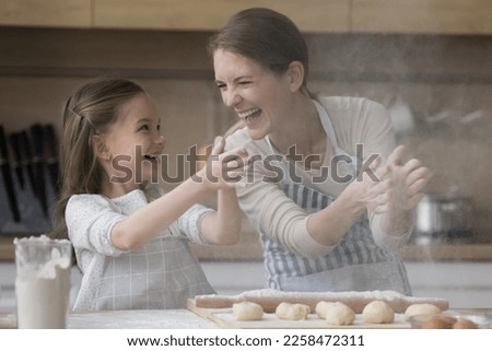 Joyful happy mother and kid girl in aprons having fun, baking in home kitchen, clapping floury hands, making powder cloud, shouting, playing, laughing at table with dough, bakery food ingredients Royalty-Free Stock Photo #2258472311