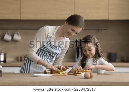 Cheerful mom and girl in aprons stacking sandwiches for lunch, smiling, laughing, enjoying culinary hobby. Daughter child helping mom to cook snacks in home kitchen together