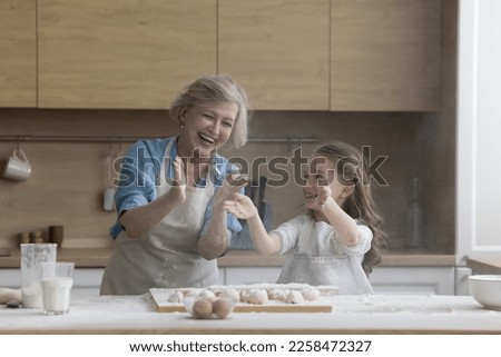 Cheerful excited grandmother and granddaughter kid having fun in home kitchen, baking, hitting floury hands over table with dough, making cloud of flour, laughing Royalty-Free Stock Photo #2258472327