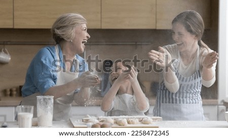 Cheerful overjoyed kid, mother and grandma having fun in kitchen, clapping floury hands over table with bakery food ingredients dough, making cloud, messy, shouting, laughing Royalty-Free Stock Photo #2258472345