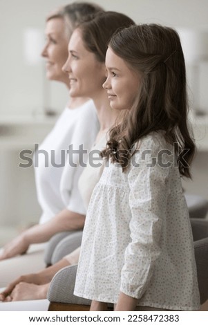 Cute pretty positive little kid girl standing by mom and grandma, looking away, smiling, thinking, dreaming. Three female family generations side portrait, vertical shot