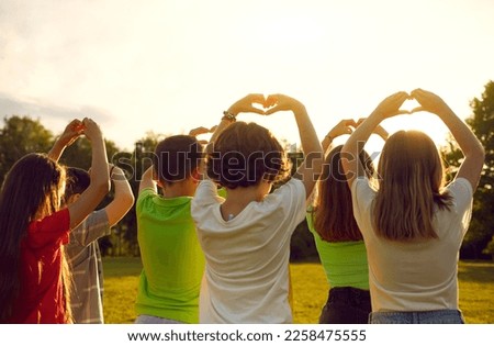 Kids wishing for peace. Back view of group of little children standing in warm evening sunlight in green park or field, putting fingers in shape of hearts and raising hands up. Love and peace concept