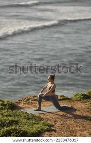 From above side view of graceful young woman with dark hair in sportswear doing stretching exercise on mat, while practicing yoga on rocky coast near wavy sea in sunlight Royalty-Free Stock Photo #2258476017