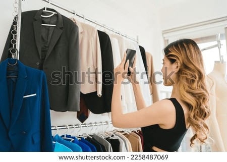A young Asian woman chooses clothes on the rail by taking a photo with smart phone for sharing on social media in a clothing store. Clothes retail shopping concept. Clipping path on a phone screen.