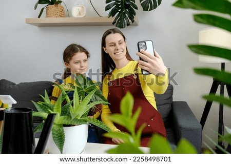 Beautiful young mother and happy little daughter record live video broadcast on modern smartphone gadget sit on sofa. Home plant breeding, gardening, housewife, online social media influencer concept