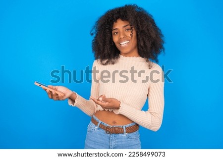 Happy pleased Young woman with afro hair style wearing crop top over blue background raises palm and holds cellphone uses high speed internet for text messaging or video calls