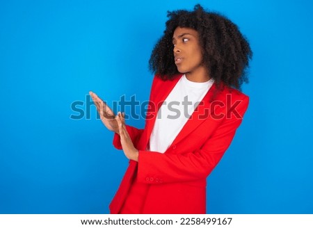 Displeased young businesswoman with afro hairstyle wearing red over blue background keeps hands towards empty space and asks not come closer sees something unpleasant