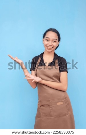 A young female barista of asian descent wearing a brown apron and black shirt presenting something to the left. Against a light blue background.