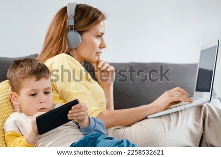 Little boy using smartphone to amuse himself while his mother is working on laptop computer. How technology affects families.