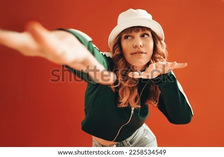 Vibrant female hipster dancing and having fun in a studio. Stylish young woman doing some moves while standing against an orange background in casual clothing.
