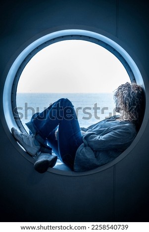Traveler passenger woman sitting inside a porthole in the boat cruise enjoying the trip and journey travel adventure alone contemplating ocean water. Concept of transport by sea and female people Royalty-Free Stock Photo #2258540739