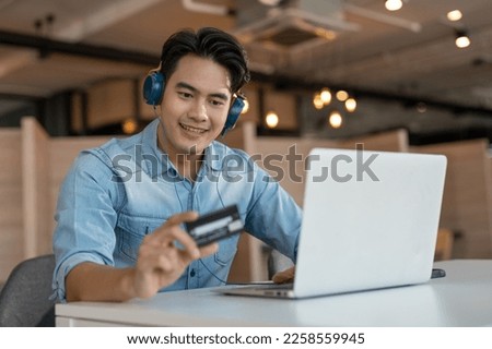 Young Asian male in blue shirt using credit card shopping online in laptop. Businessman making payment purchasing on internet.