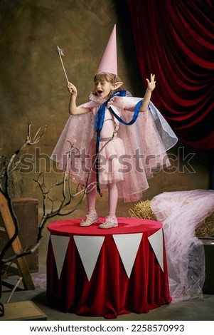 Little performer. Beautiful cute girl, child in image of magical elf posing in pink dress over vintage circus background. Making magic. Retro circus performance. Dreams, art, vintage style, childhood
