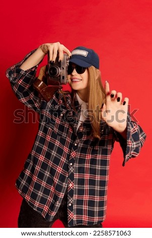 Photo in moment. Beautiful young woman in checkered shirt, cap and sunglasses posing with photo camera over red studio background. Concept of youth, beauty, lifestyle, emotions, facial expression. Ad