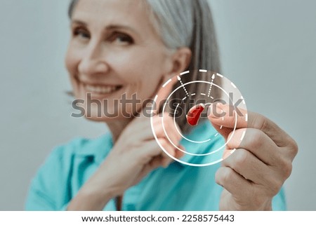 Mature woman holding BTE hearing aid in hand on foreground and points to her ear, concept. Hearing loss treatment Royalty-Free Stock Photo #2258575443