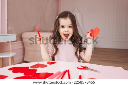 a little girl makes Valentine's Day cards using colored paper, scissors and pencil, sitting at a table in a cozy room. Royalty-Free Stock Photo #2258578465