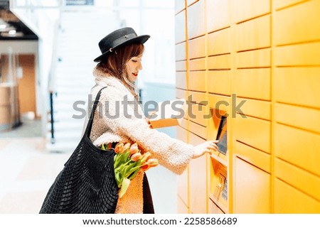 Fashion woman with box using modern postal automatic mail terminal with self service device for pickup or refund an order. Electronic locker for storing parcels. Online shopping. Selective focus Royalty-Free Stock Photo #2258586689