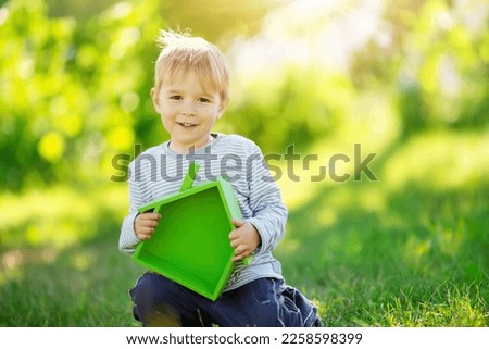Smiling child sitting on the spring meadow with model of the green house in his hands. Concept of the real estate, rental and buying of a new apartment.