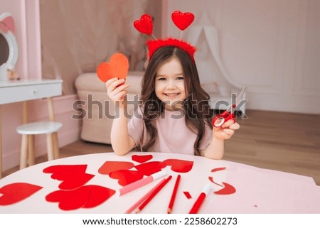 a little girl makes Valentine's Day cards using colored paper, scissors and pencil, sitting at a table in a cozy room. Royalty-Free Stock Photo #2258602273