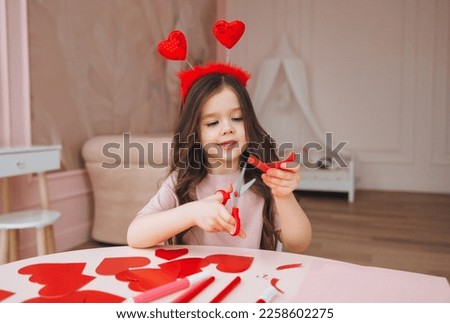 a little girl makes Valentine's Day cards using colored paper, scissors and pencil, sitting at a table in a cozy room.
