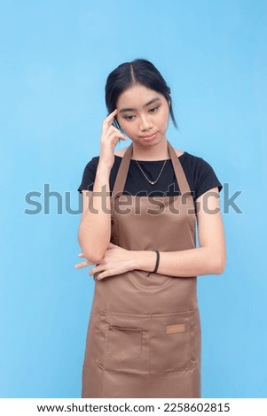 A cute and pretty asian barista or waitress in deep thought, worried about her job. Isolated on a light blue background. Royalty-Free Stock Photo #2258602815