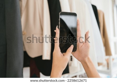 A close up woman chooses clothes on the rail by taking a photo with smart phone for sharing on social media in a clothing store. Clothes retail shopping concept. Clipping path on a phone screen.