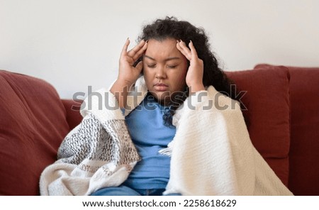 Sick unhappy overweight brunette curly young woman covered in warm blanket sitting on couch at home, touching head, suffering from headache during coronavirus or flu, copy space Royalty-Free Stock Photo #2258618629