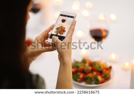 Unrecognizable brunette woman taking photo of festive table with strawberry, wine and candles while St. Valentines Day celebration or romantic date with her boyfriend, using brand new phone, cropped