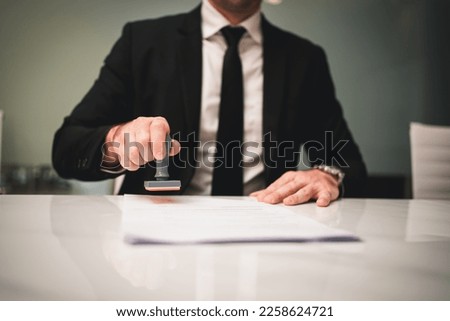 Businessman hand stamping an approved stamp on a text document at a table with a contract form paper in the background.