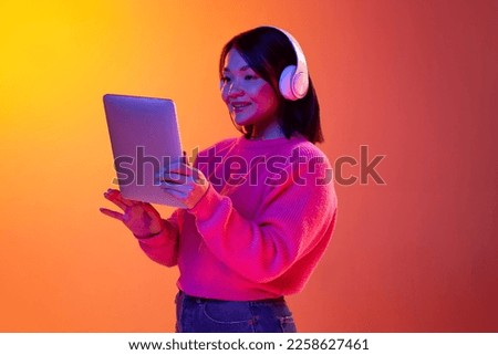 Online lessons. Young girl, student in headphones looking on tablet over gradient orange background in neon light. Concept of emotions, facial expression, youth, inspiration, sales, ad