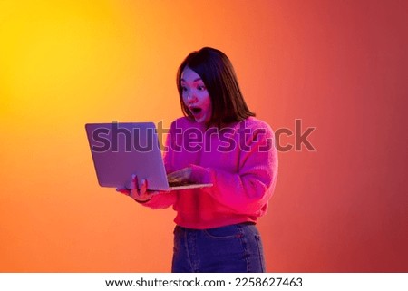 Shocked news. Young girl in pink sweater emotionally looking on laptop over gradient orange background in neon light. Freelancer. Concept of emotions, facial expression, youth, inspiration, sales, ad